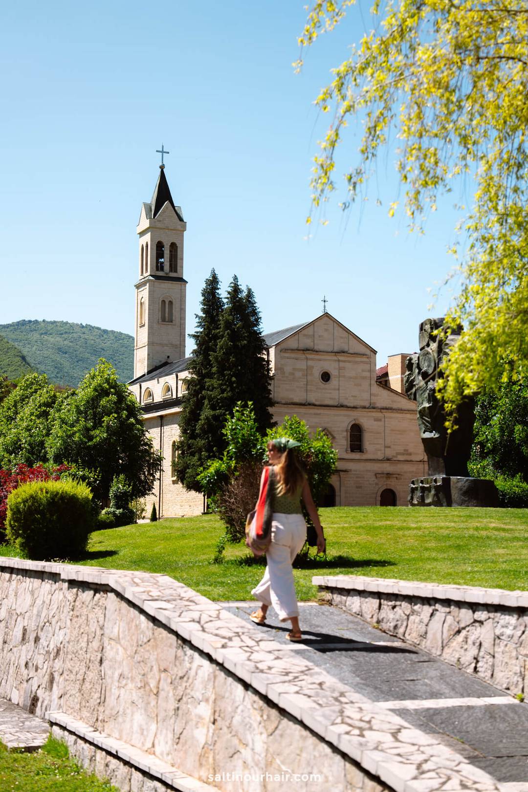 A woman walks across a stone bridge toward a church with a tall bell tower, surrounded by green trees and grass under a clear blue sky&mdash;a perfect stop on any Bosnia itinerary.