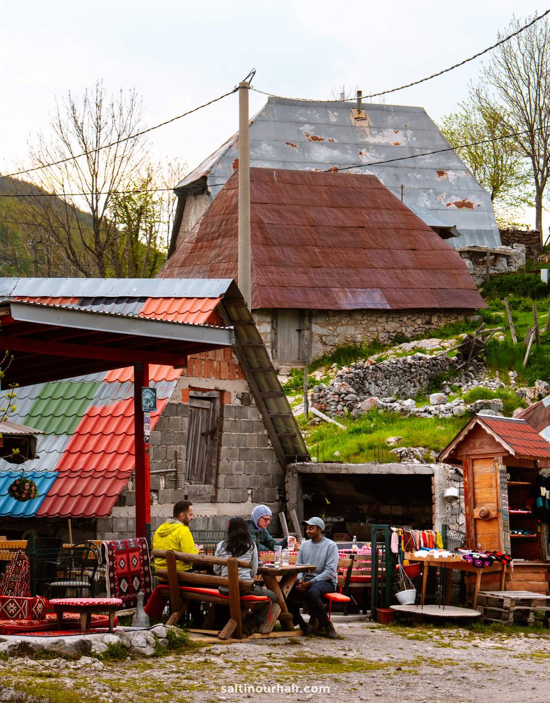 Four people sit at an outdoor table in a rustic village setting&mdash;colorful roofs, wooden buildings, and traditional crafts on display&mdash;perfect for anyone exploring a Bosnia itinerary.