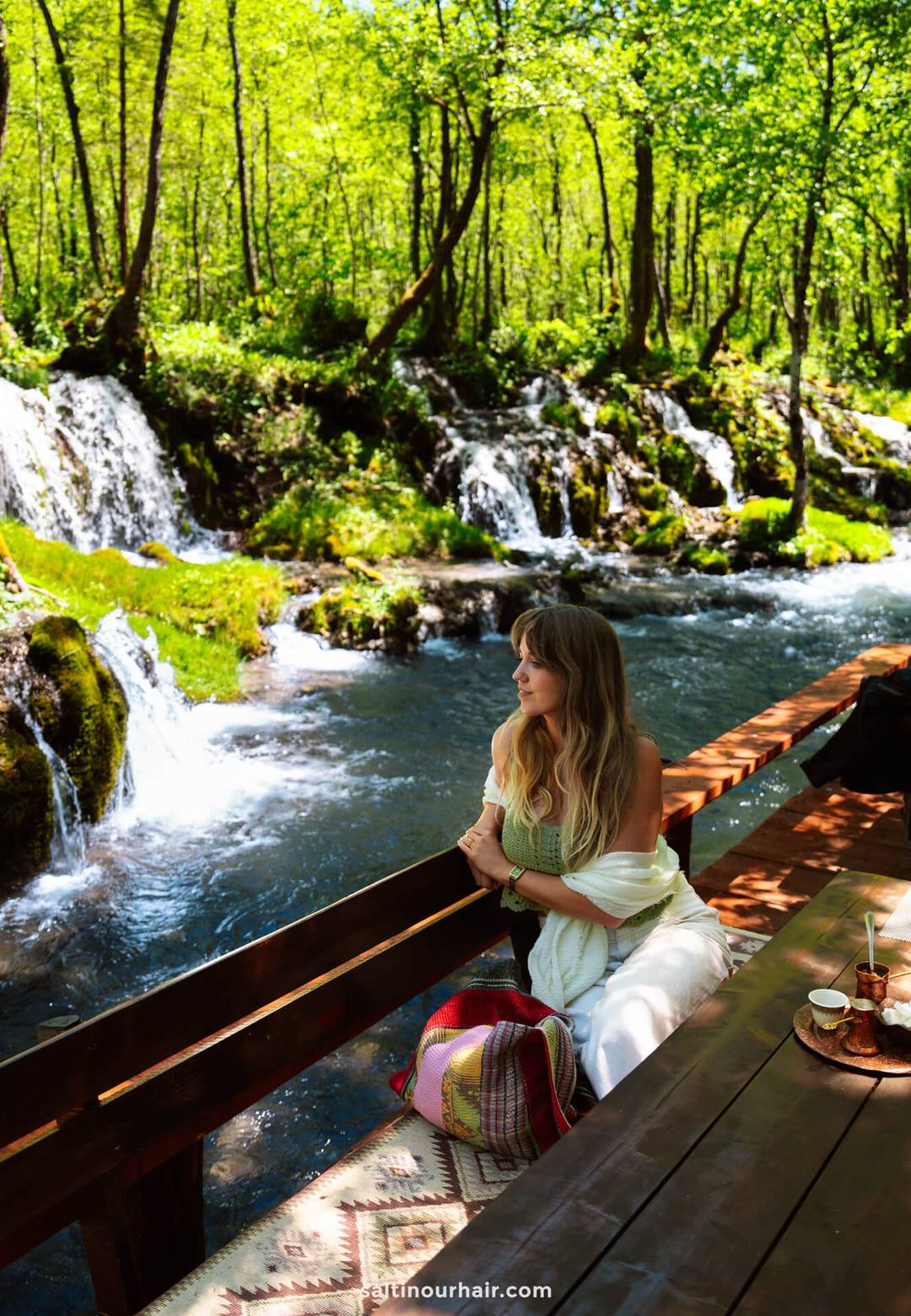 A woman sits at a wooden table by a river with waterfalls, surrounded by lush green trees&mdash;an idyllic moment that could inspire any Bosnia itinerary. Tea cups and a small teapot rest on the table, completing the tranquil scene.