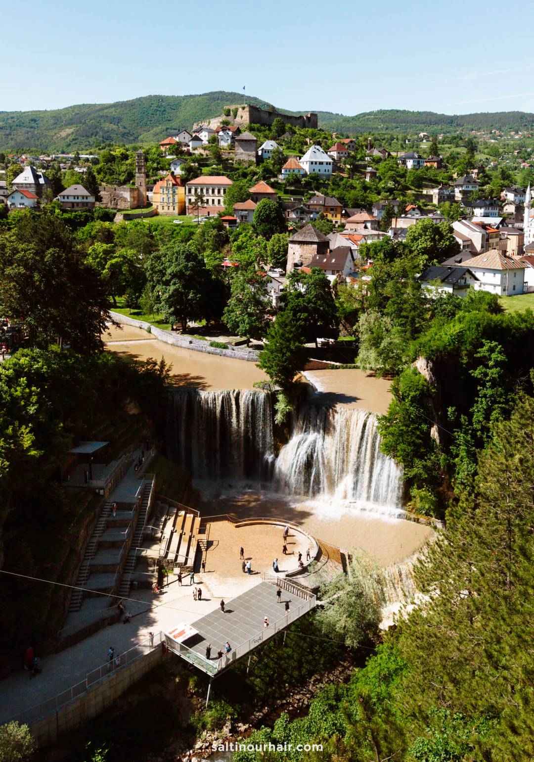 Aerial view of a waterfall with a viewing platform below, surrounded by trees and a town with colorful buildings and hills in the background&mdash;an essential stop on any Bosnia itinerary.