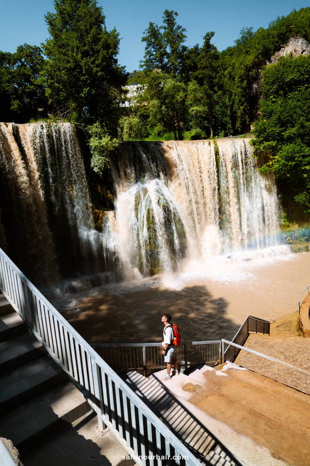 A person with a red backpack stands on stairs overlooking a large waterfall, a perfect stop on any Bosnia itinerary, surrounded by trees under a clear blue sky.