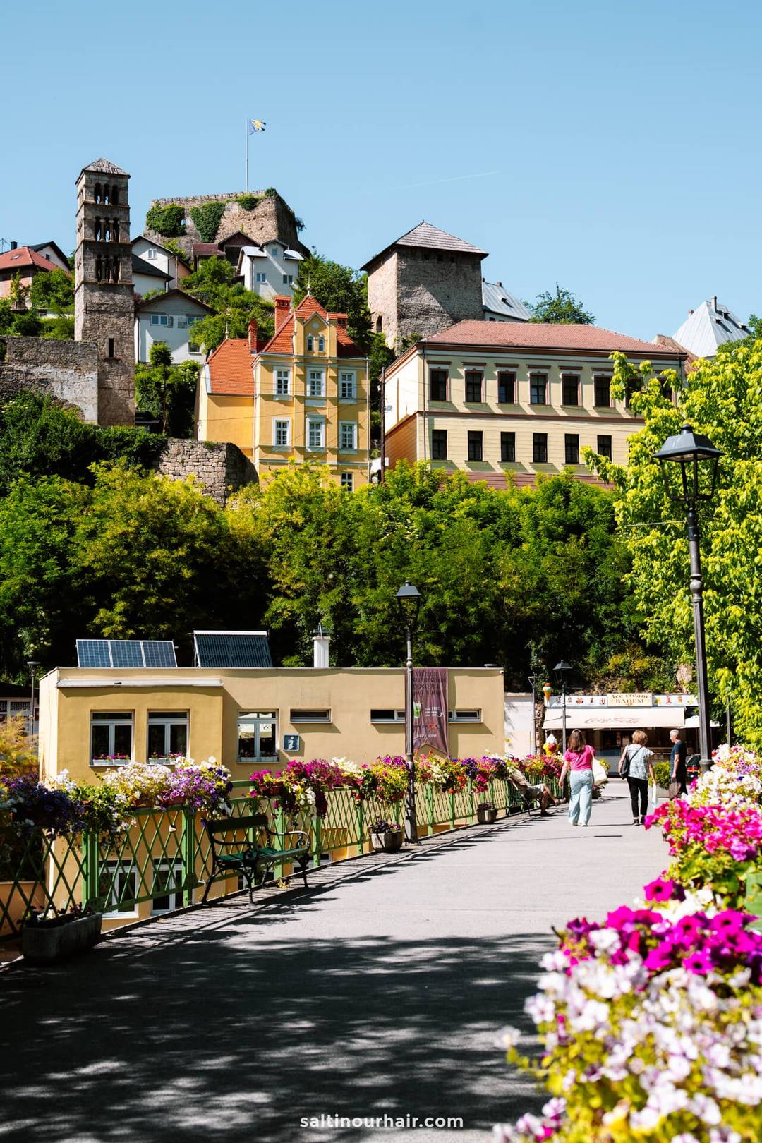 A walkway lined with colorful flowers leads to historic buildings and a hilltop stone tower under a clear blue sky&mdash;an iconic stop on any Bosnia itinerary, where people stroll along the vibrant path.