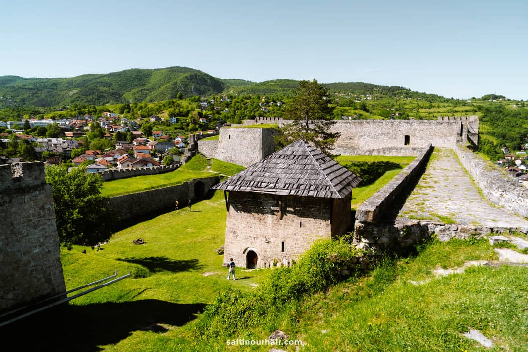 A historic stone fortress with towers and a wooden-roofed building sits on a grassy hill, offering a must-see view for any Bosnia itinerary, overlooking a town and forested mountains under a clear blue sky.