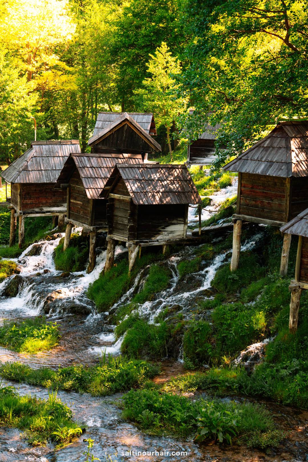 Several small wooden huts are built on stilts above a stream with cascading water, surrounded by lush green trees and grass&mdash;a hidden gem worth adding to any Bosnia itinerary.