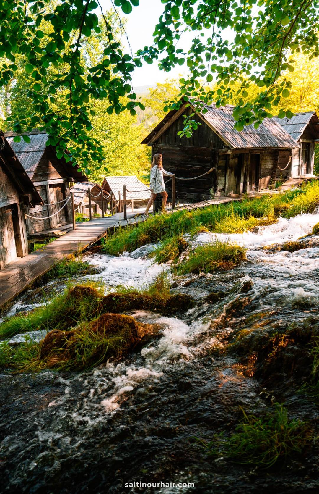A person walks on a wooden path beside old wooden cabins and a rushing stream, surrounded by green trees and sunlight&mdash;an idyllic stop on any Bosnia itinerary.