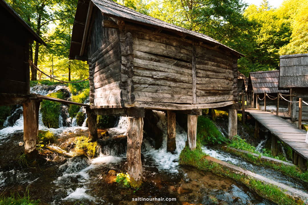 Wooden water mill cabins stand on stilts above flowing streams and small waterfalls, surrounded by green trees and a wooden walkway in daylight&mdash;an enchanting stop on any Bosnia itinerary.