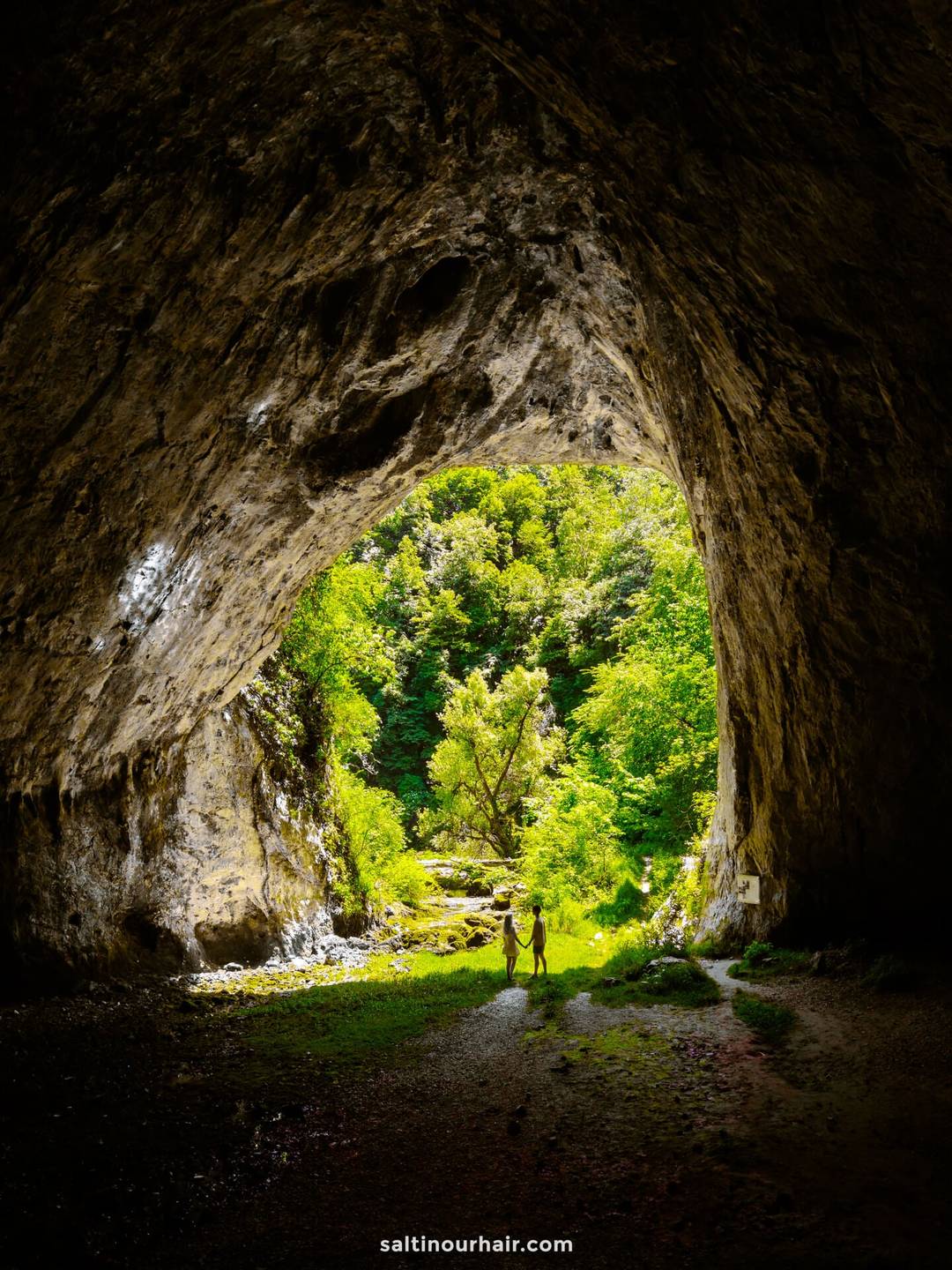 A view from inside a large cave looking out toward greenery and trees, with two people standing near the cave entrance&mdash;an unforgettable stop on any Bosnia itinerary.
