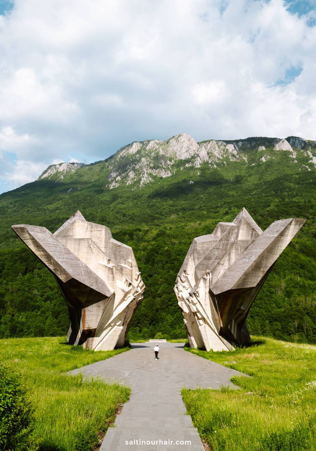 A person walks toward a large, angular concrete monument&mdash;an essential stop on any Bosnia itinerary&mdash;set against a backdrop of green hills and mountains under a partly cloudy sky.