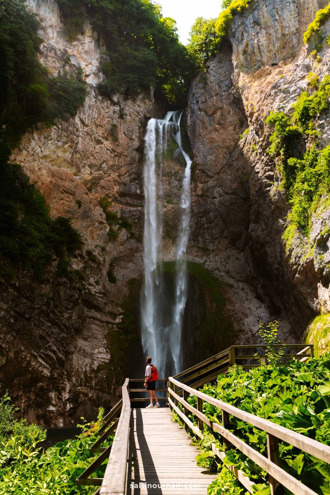 A person stands on a wooden walkway facing a tall waterfall cascading down a rocky cliff surrounded by greenery&mdash;a stunning highlight for any Bosnia itinerary.