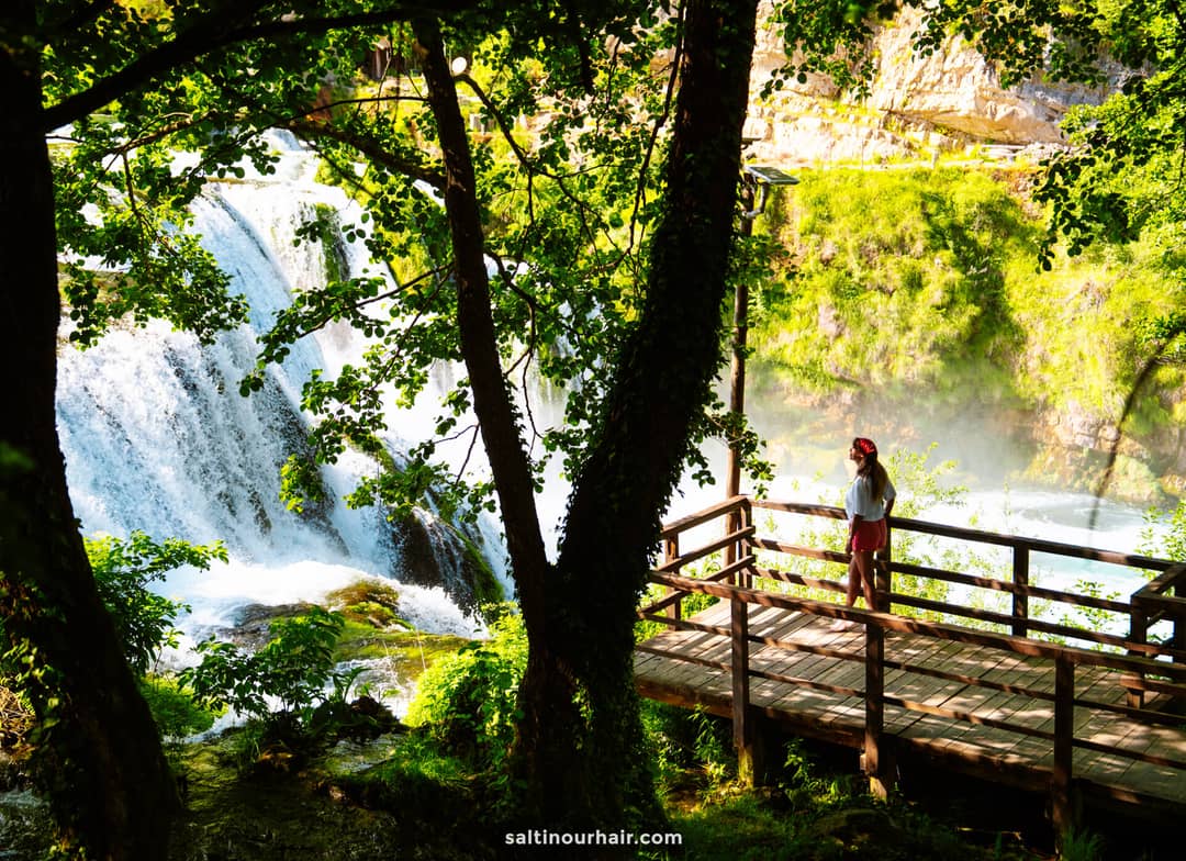 A person stands on a wooden platform overlooking a large waterfall, surrounded by green trees and sunlight&mdash;an unforgettable stop on any Bosnia itinerary.