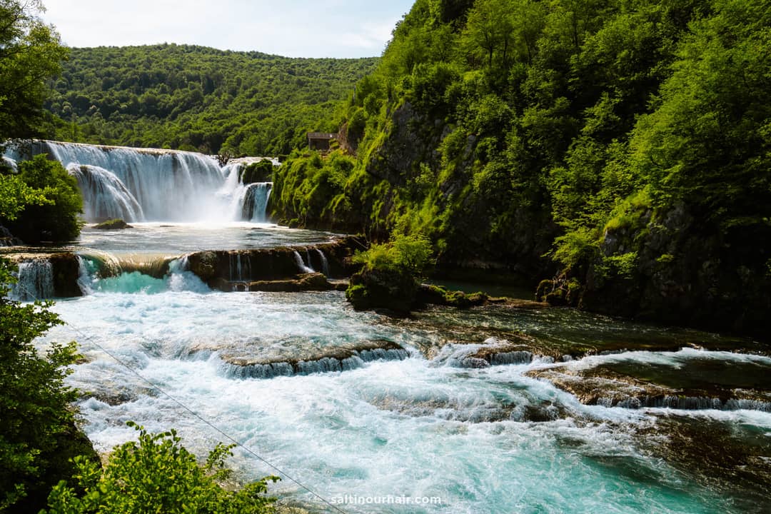 A wide river flows over a multi-tiered waterfall, surrounded by dense green trees and hills under a partly cloudy sky&mdash;a must-see stop on any Bosnia itinerary.