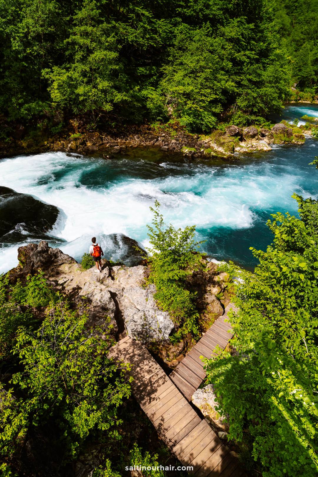 A person in a red shirt stands on a rocky outcrop beside a fast-flowing blue river, surrounded by dense green forest and a wooden footbridge&mdash;a perfect scene for any Bosnia itinerary.