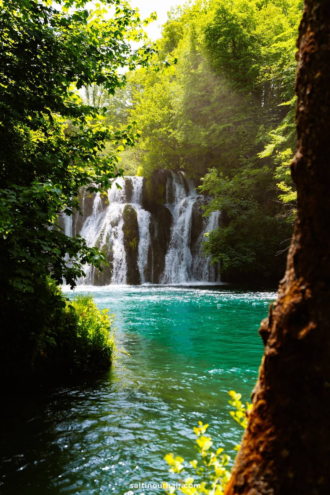 A waterfall cascades into a turquoise pool surrounded by lush green trees, with sunlight filtering through the foliage&mdash;an unforgettable highlight for any Bosnia itinerary.