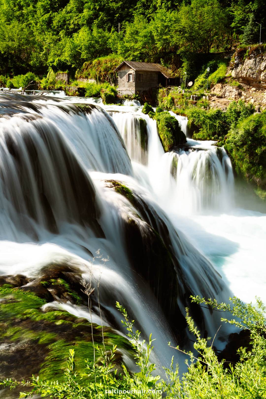 A wooden cabin sits near large, cascading waterfalls surrounded by lush greenery and rocks under bright daylight&mdash;a tranquil highlight for any Bosnia itinerary.