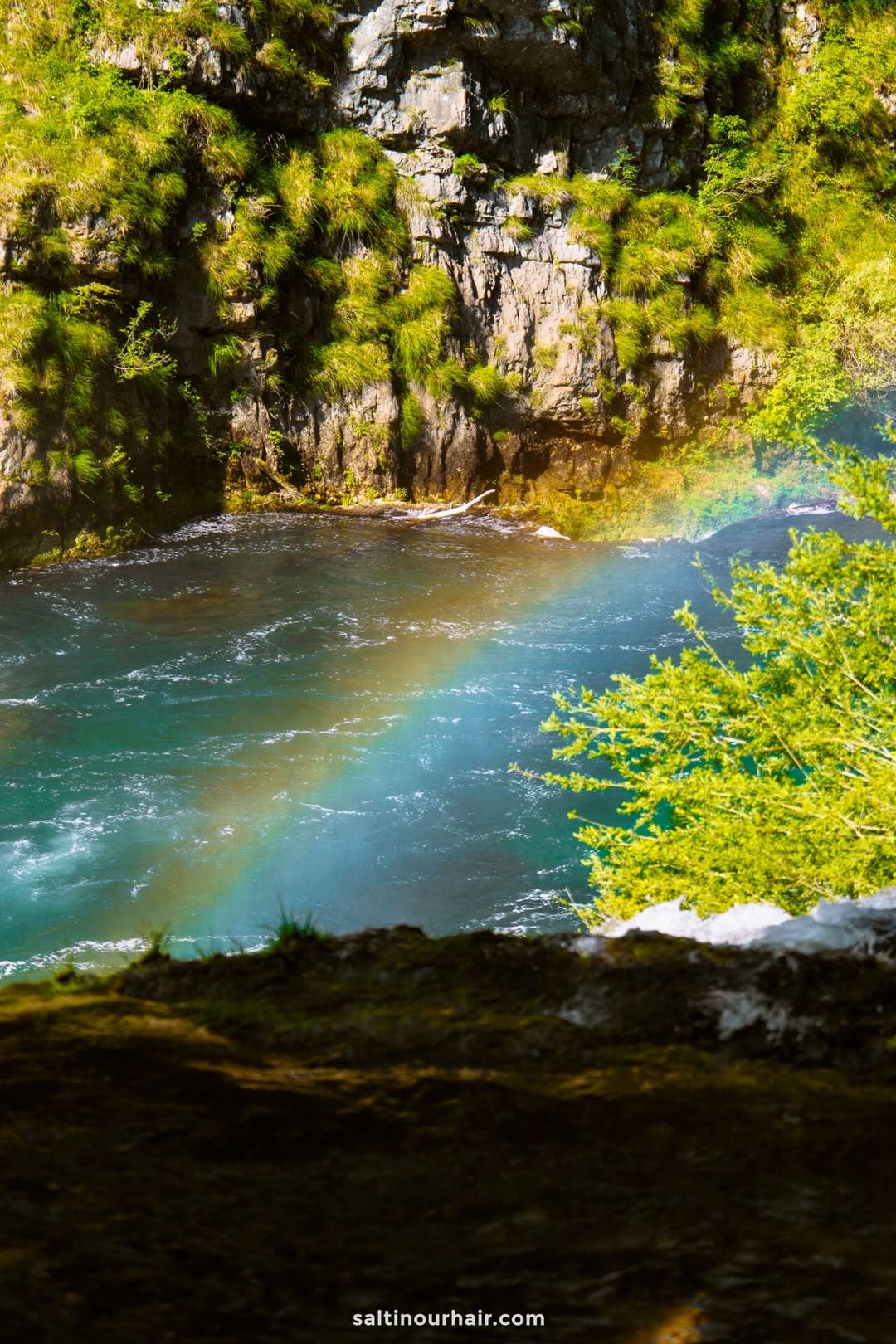 A rainbow appears over a blue river with rocky cliffs and green vegetation in sunlight&mdash;a perfect view to add to your Bosnia itinerary.
