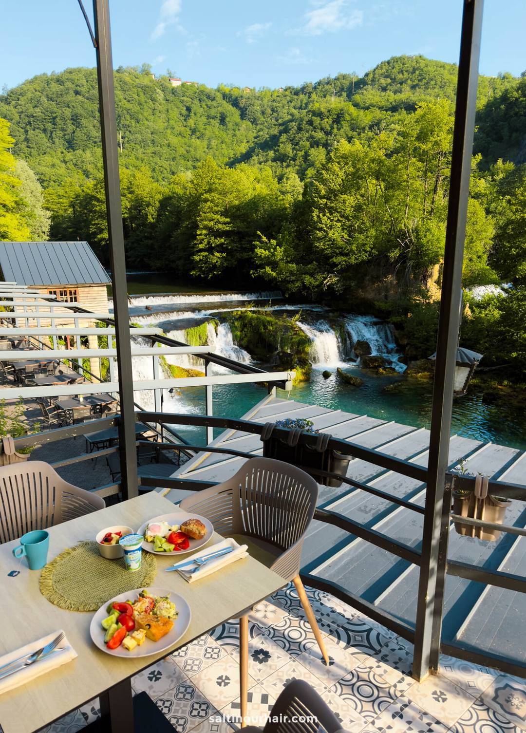 Outdoor dining area overlooking a small waterfall and lush green trees, with plates of food and drinks set on a table&mdash;an ideal spot to include in your Bosnia itinerary.