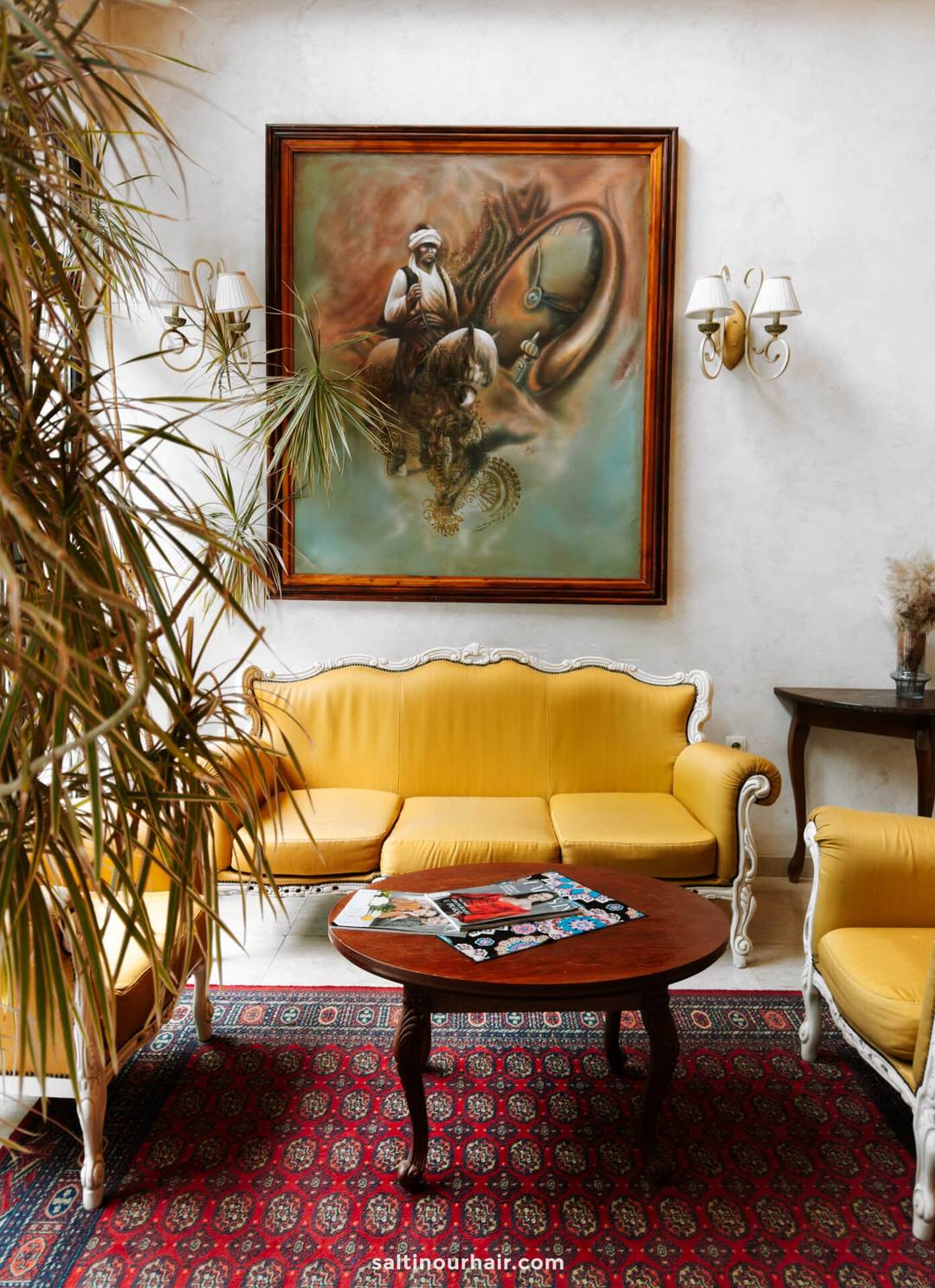 A living room with yellow upholstered furniture, a wooden table, a red patterned rug, and a large framed painting of a man with a horse offers inspiration for any Bosnia itinerary.