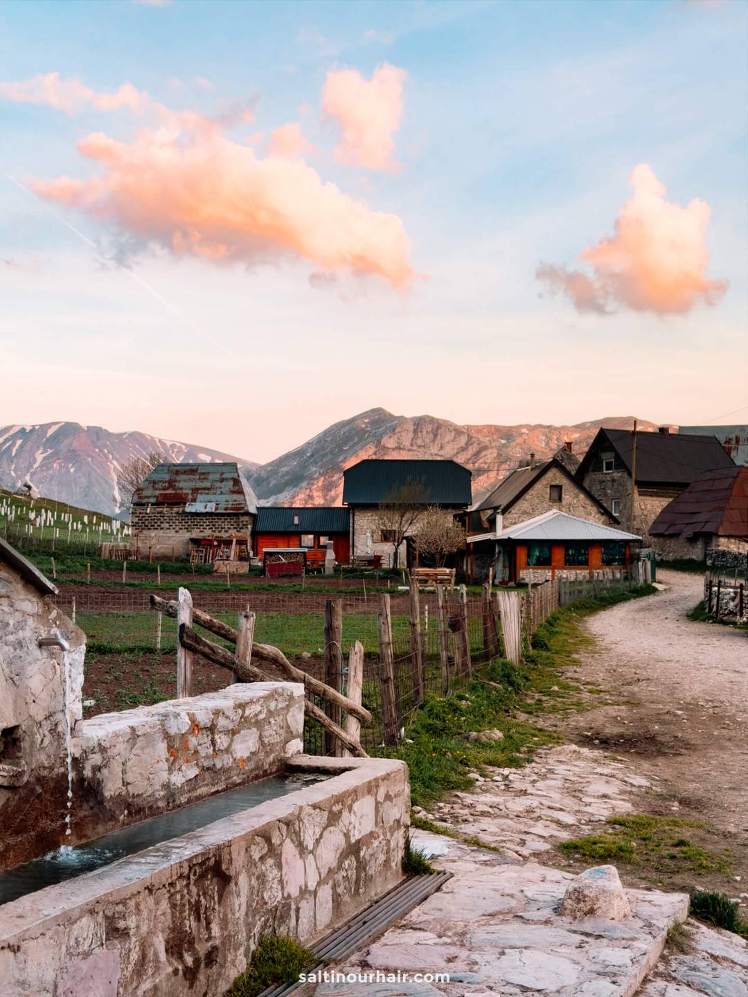A rural village, perfect for a Bosnia itinerary, with houses, a dirt path, wooden fences, and a stone water trough, set against mountains under a sky with pink clouds at sunset.