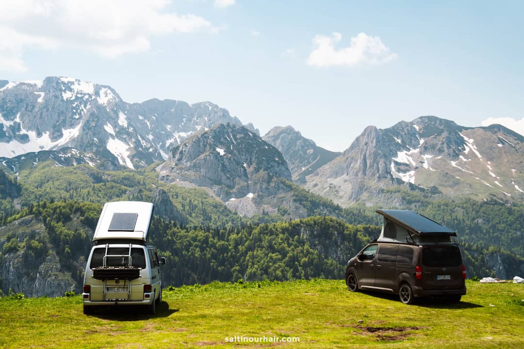 Two camper vans parked on grass with open rooftop tents overlook a mountain range with snowy patches and green forest&mdash;perfect for planning your next Bosnia itinerary.