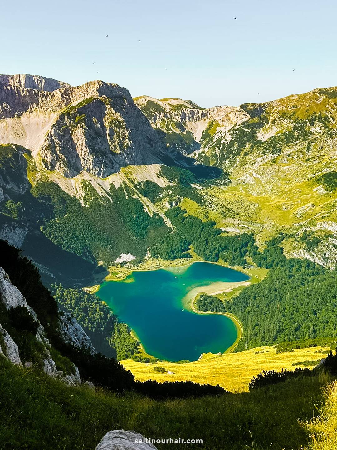 the turquoise Trnovačko Lake surrounded by green forests and rocky mountains under a clear sky, viewed from above&mdash;an unforgettable hike on any Bosnia itinerary.