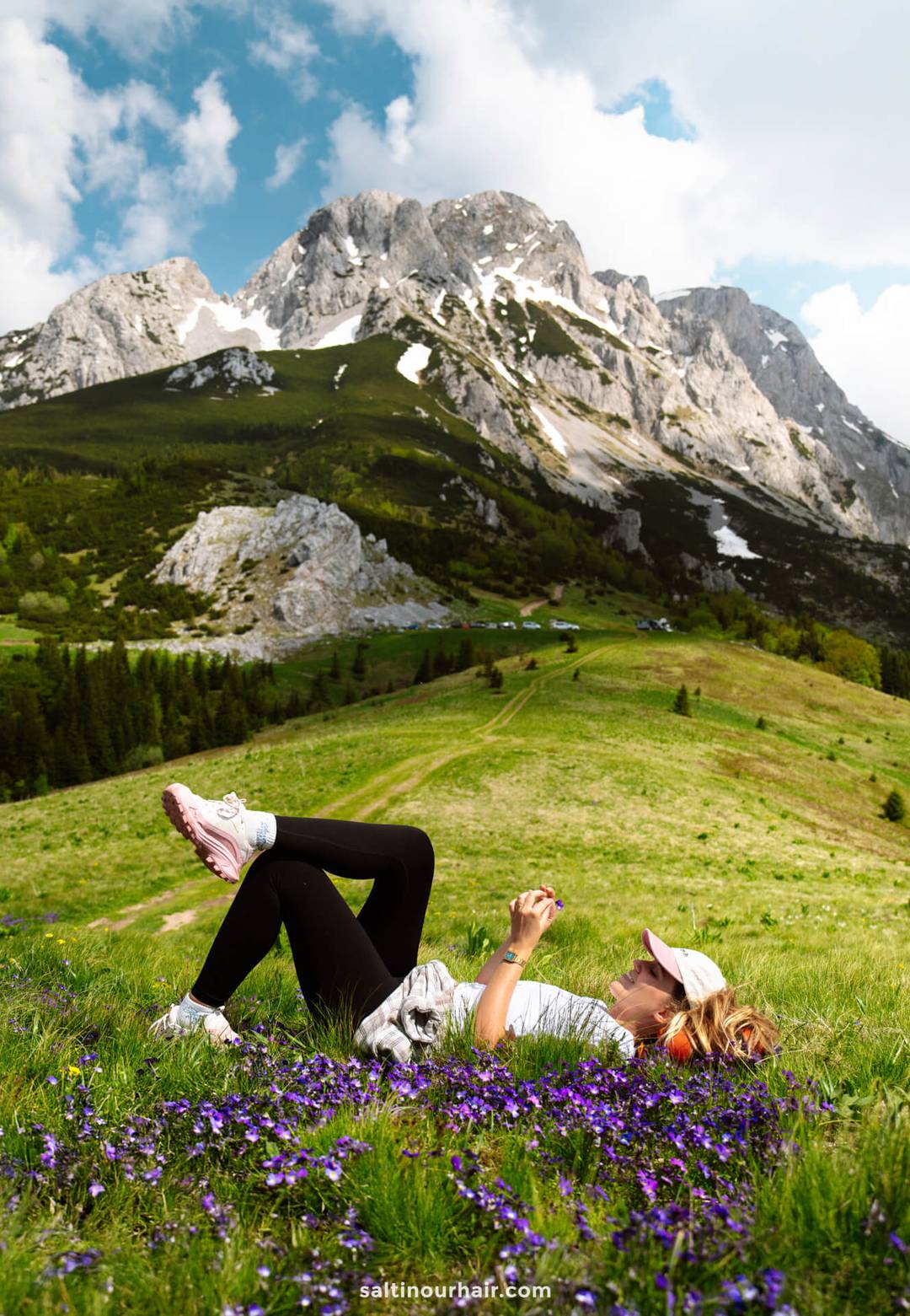 A woman lies on grass among purple flowers in a mountain meadow, planning her Bosnia itinerary on her phone, with rocky peaks and clouds in the background.