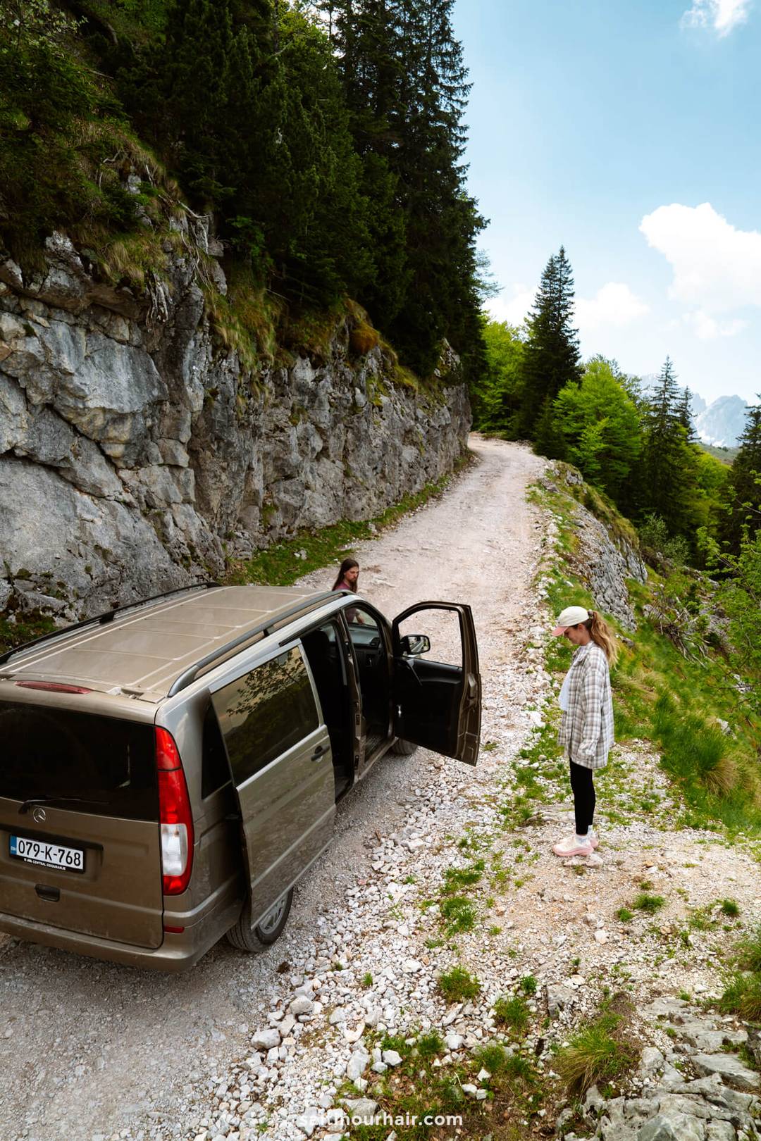 A brown van is parked on a rocky mountain road&mdash;a perfect stop on your Bosnia itinerary&mdash;with one person standing outside and another getting out, surrounded by trees and cliffs.