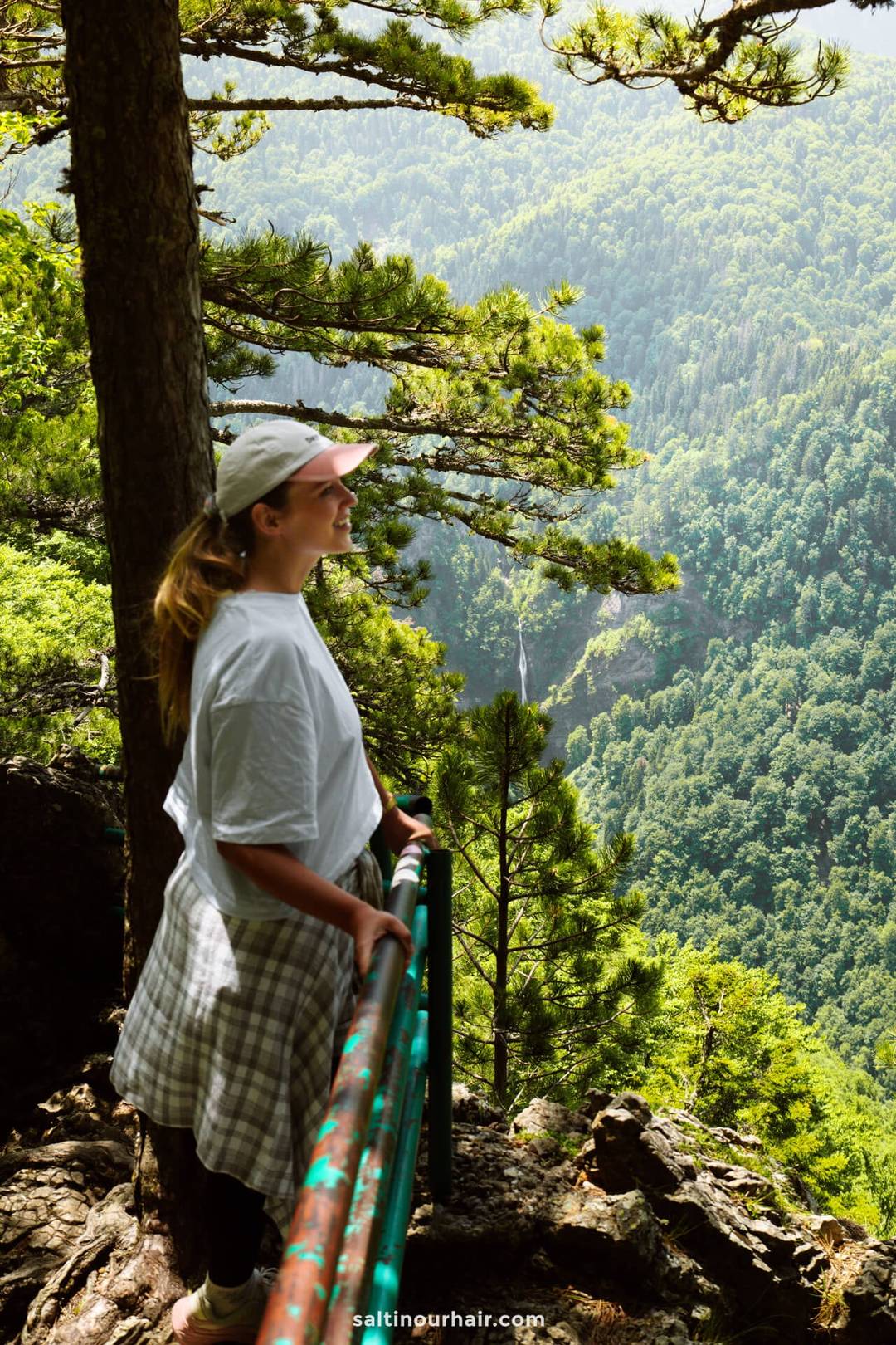 A person wearing a white cap and shirt stands by a green railing on a forested mountain overlook, perhaps considering their next stop on a Bosnia itinerary, while gazing at the distant valley.