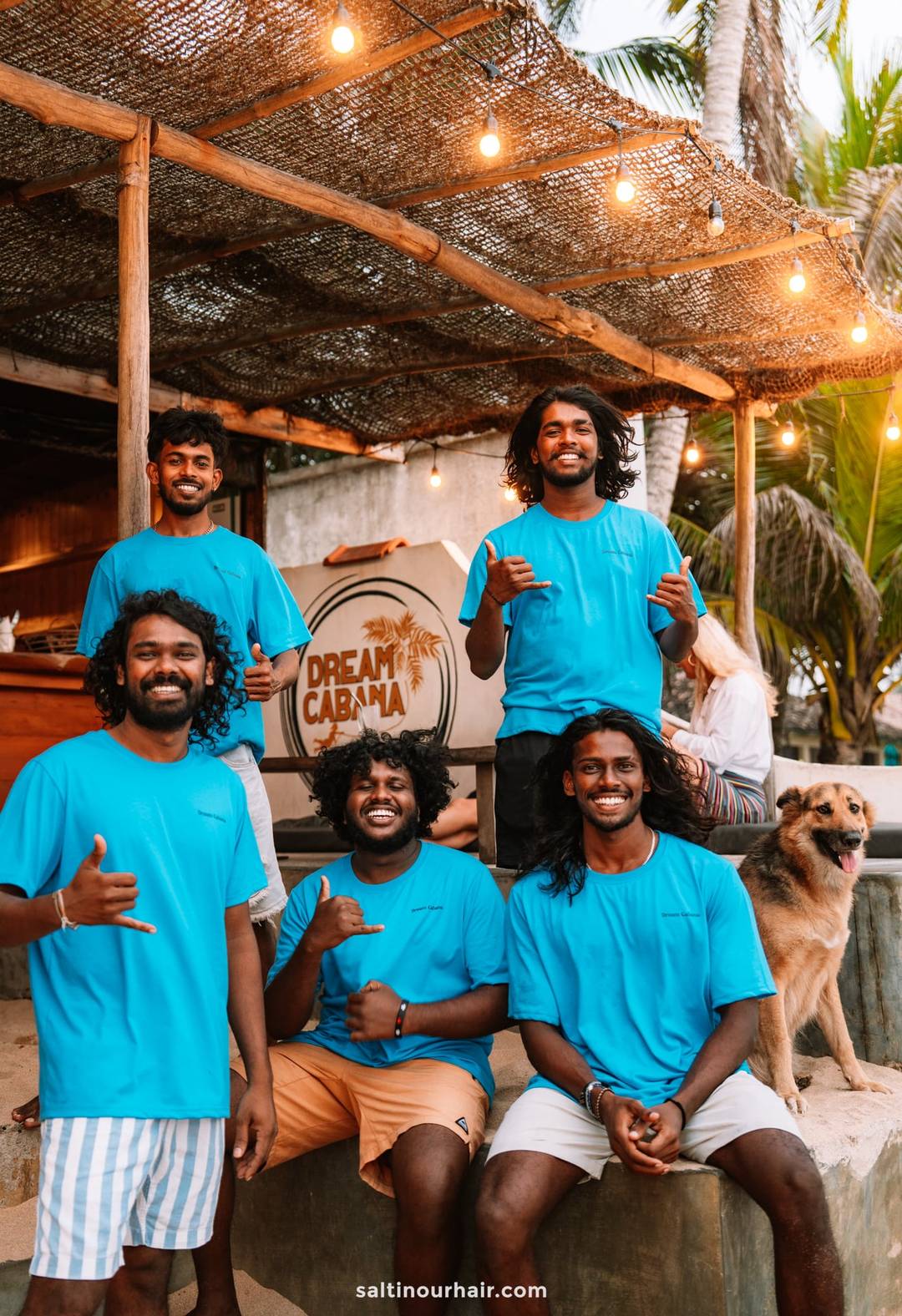 Five smiling Dream Cabana staff in matching blue T-shirts posing under string lights at the beach bar in Ahangama, Sri Lanka.