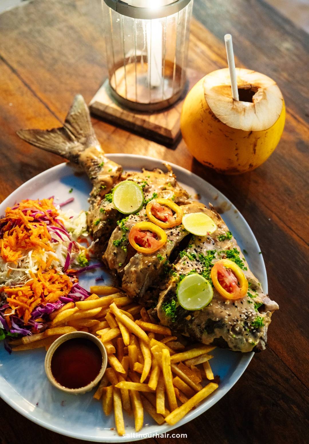 Whole grilled fish topped with lime slices, tomatoes, and herbs, served with fries, coleslaw, and a fresh coconut drink in Ahangama.