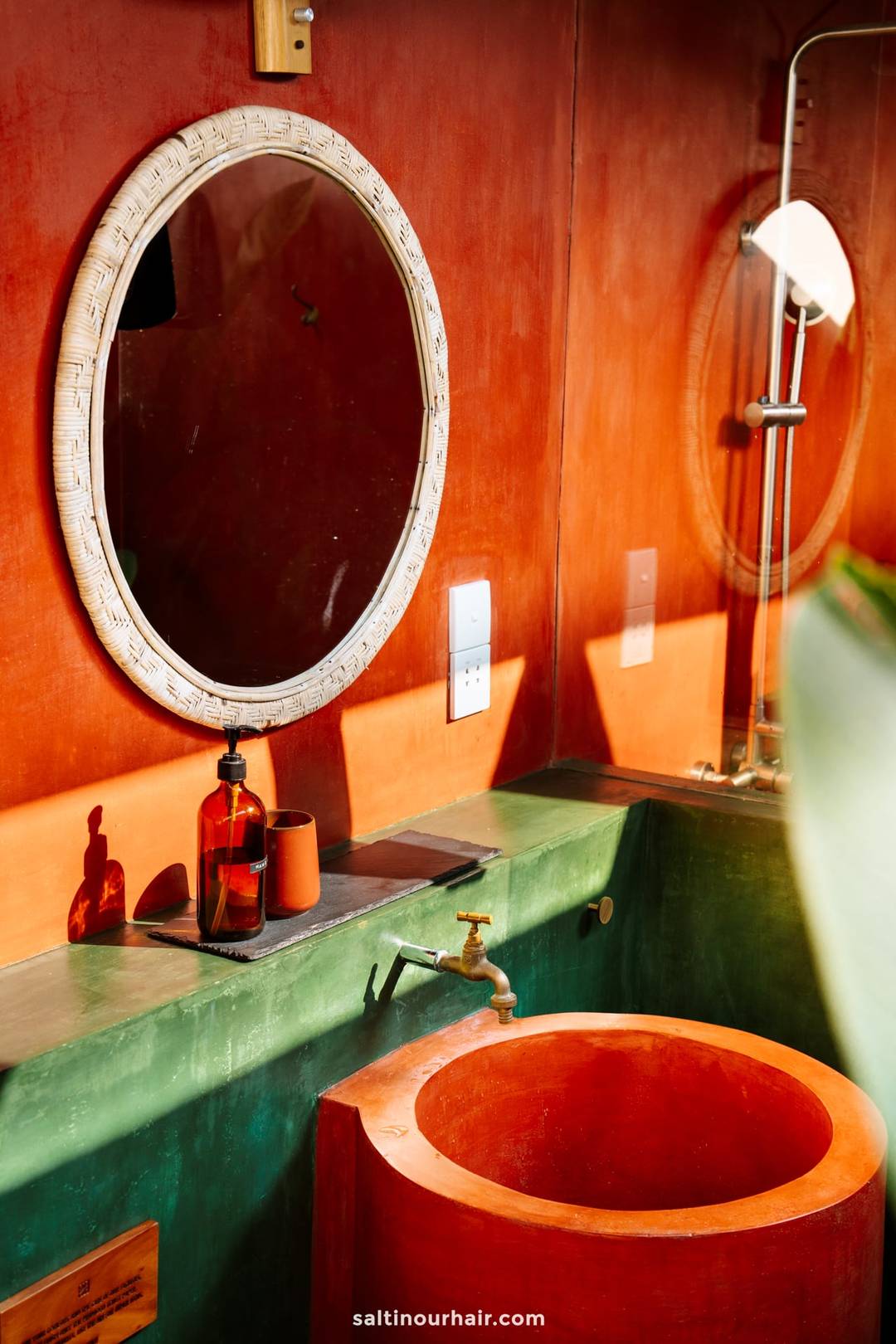 Colorful boutique bathroom with a round rattan mirror, terracotta sink, brass faucet, and green concrete vanity in Ahangama.