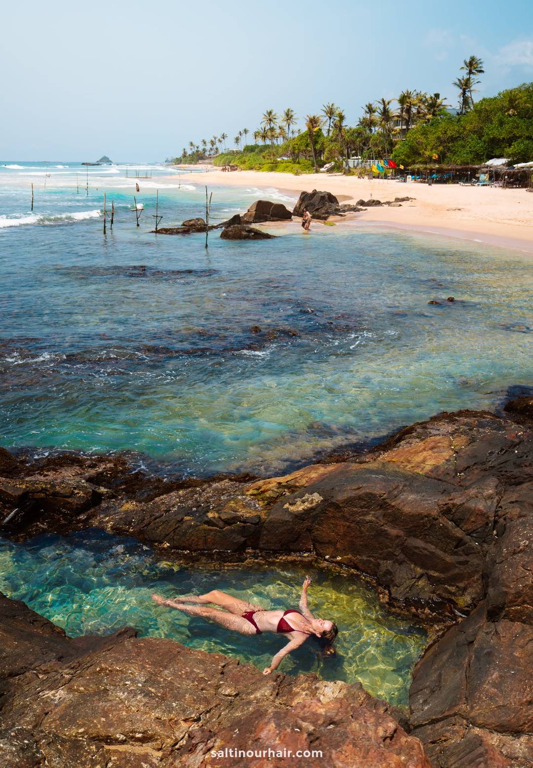 Woman floating in a rocky tidal pool at Coconut Beach in Ahangama, Sri Lanka, with stilt fishermen visible beyond.