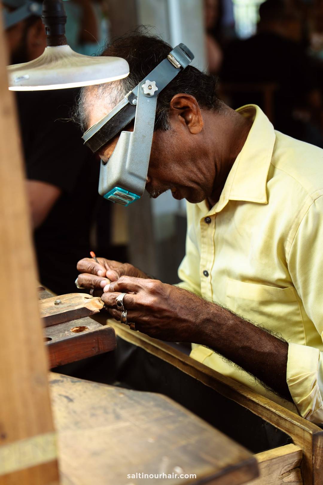 Sri Lankan jeweler wearing magnifying headset carefully crafting jewelry at a wooden workbench in Ahangama.