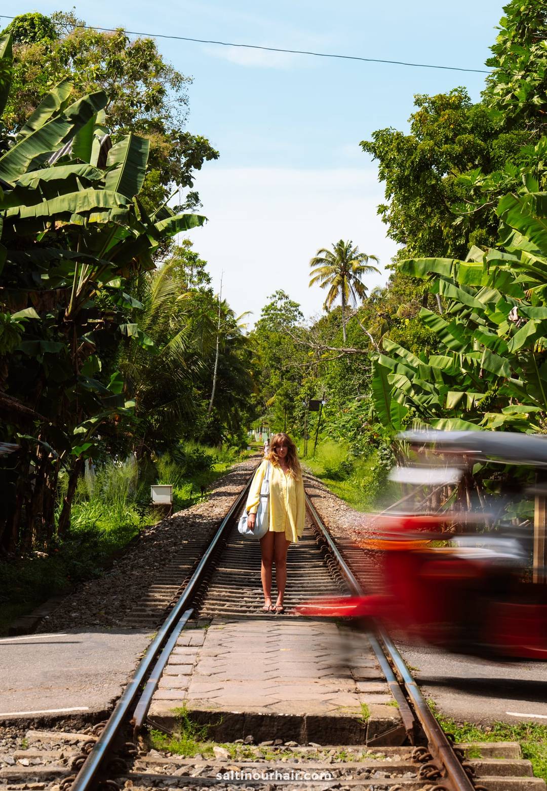 Woman in yellow dress standing on Ahangama railway tracks as a red tuk-tuk speeds past, surrounded by tropical jungle.