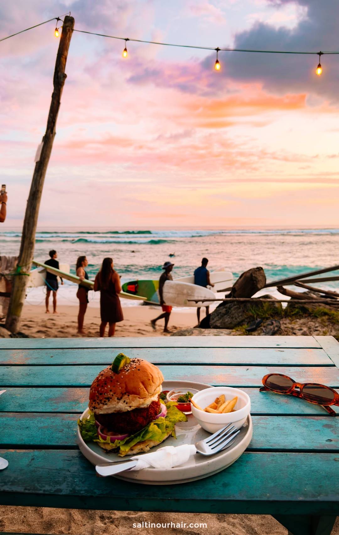 Burger with lettuce and avocado served beachside at Ahangama beach caf&eacute;, surfers carrying boards at sunset in background.