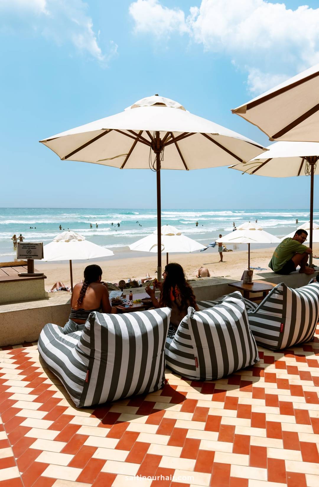 Two women dining on gray and white striped bean bag chairs on a red geometric-tiled terrace overlooking Ahangama beach.