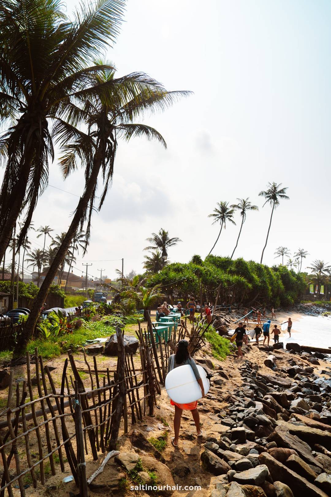 Surfer carrying a white board toward Ahangama beach, Sri Lanka, past a rustic wooden fence and palm trees.