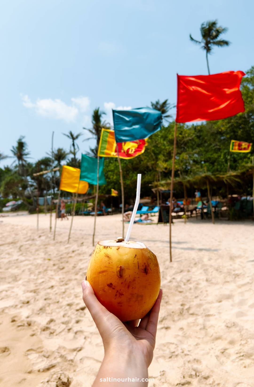 Fresh king coconut with a straw held up at Coconut Beach in Ahangama, Sri Lanka, with colorful flags flying.