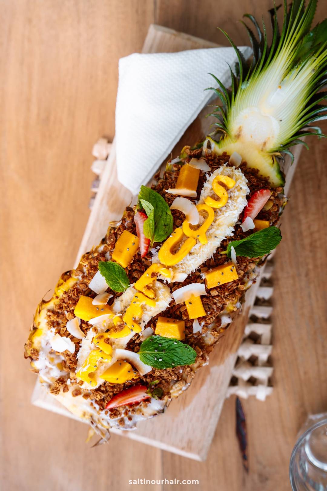 Tropical pineapple boat smoothie bowl topped with granola, mango chunks, strawberries, coconut flakes, and fresh mint leaves.