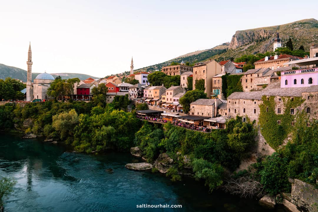 Mostar old town skyline with Koski Mehmed Pasha Mosque, riverside restaurants, and stone buildings above the Neretva River.