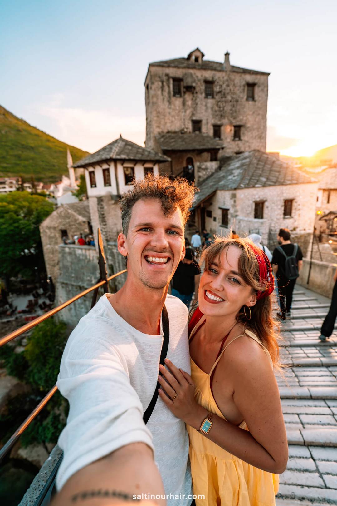 A smiling couple takes a selfie on a stone bridge at sunset, with historic stone buildings and hills in the background&mdash;capturing one of the must-see things to do in Mostar, Bosnia.