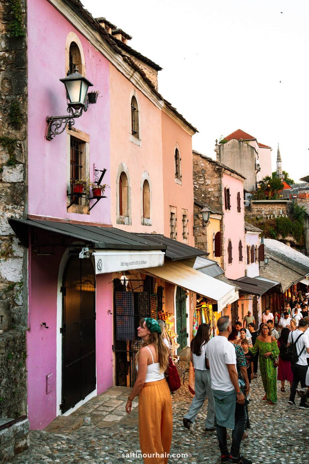 A street scene with people walking past colorful pink buildings, stone facades, and vibrant shops in a historic area at sunset&mdash;one of the charming things to do in Mostar, Bosnia.