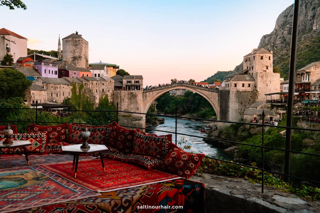 Outdoor seating area with patterned rugs and cushions overlooking the historic Stari Most bridge at sunset&mdash;one of the top things to do in Mostar, Bosnia and Herzegovina.