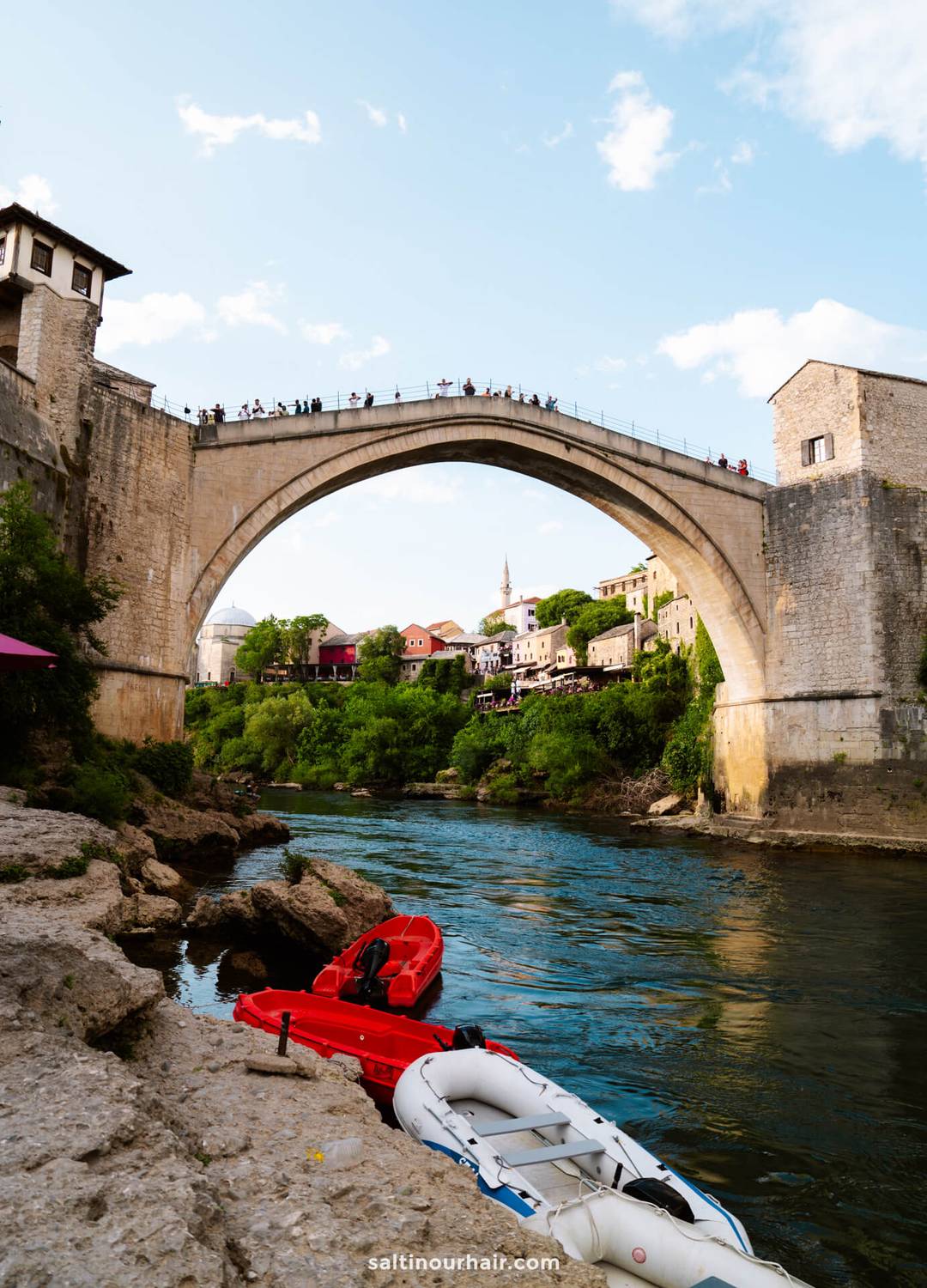 A stone arched bridge spans a river, with people standing on top&mdash;a classic scene among the things to do in Mostar, Bosnia. Red and white kayaks rest on the rocky shore, while buildings and greenery line the riverbanks.