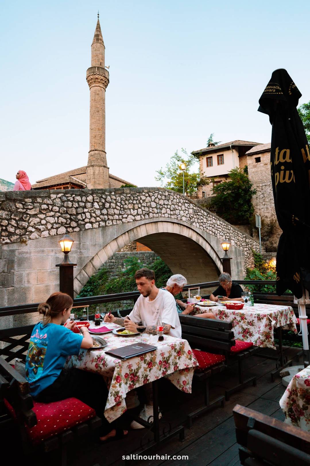 Outdoor restaurant terrace with floral tablecloths beside Crooked Bridge and mosque minaret in Mostar, Bosnia.