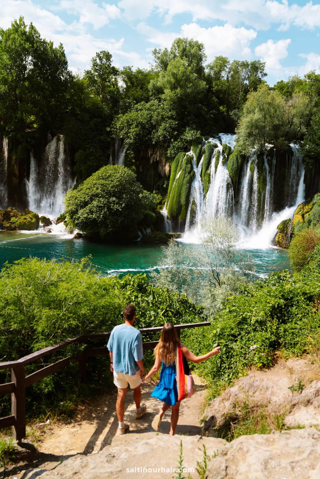 A man and woman walk down a path surrounded by greenery toward a scenic waterfall and blue water under a partly cloudy sky&mdash;one of the top things to do in Mostar, Bosnia.
