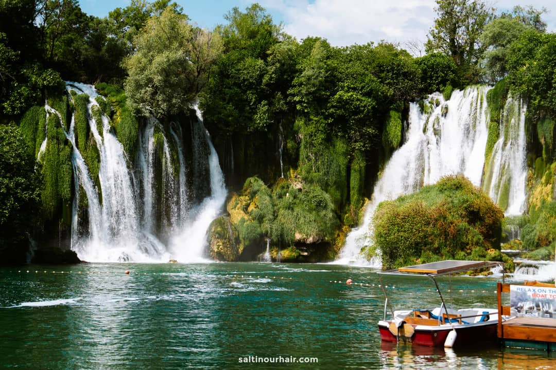 Kravica Waterfalls cascading over moss-covered rocks into turquoise water, with small wooden boats docked in the foreground.