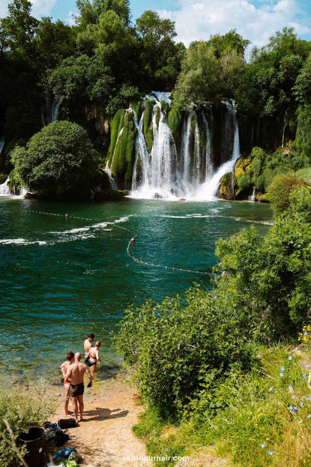 People stand at the edge of a lush, green lake with a large waterfall in the background on a sunny day&mdash;one of the unforgettable things to do in Mostar, Bosnia.
