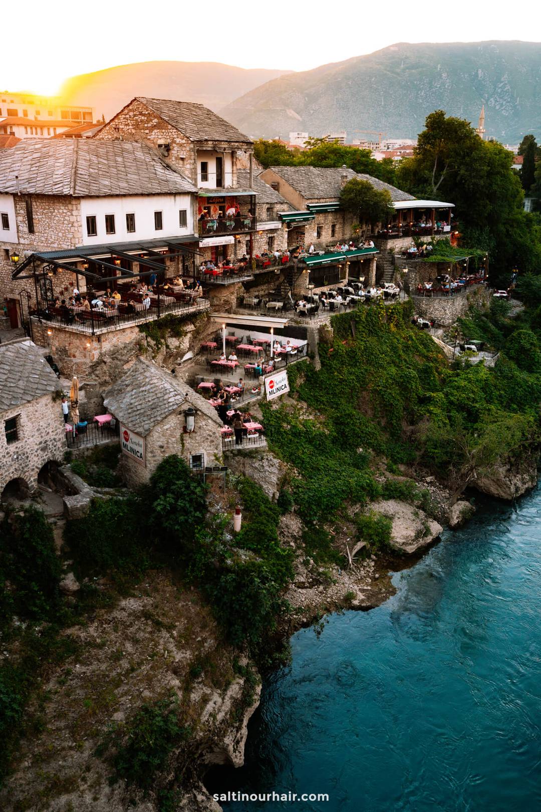 Stone buildings and cliffside cafes overlook a river with clear blue water; lush greenery climbs the rocky bank, and mountains rise in the background&mdash;just some of the things to do in Mostar, Bosnia.