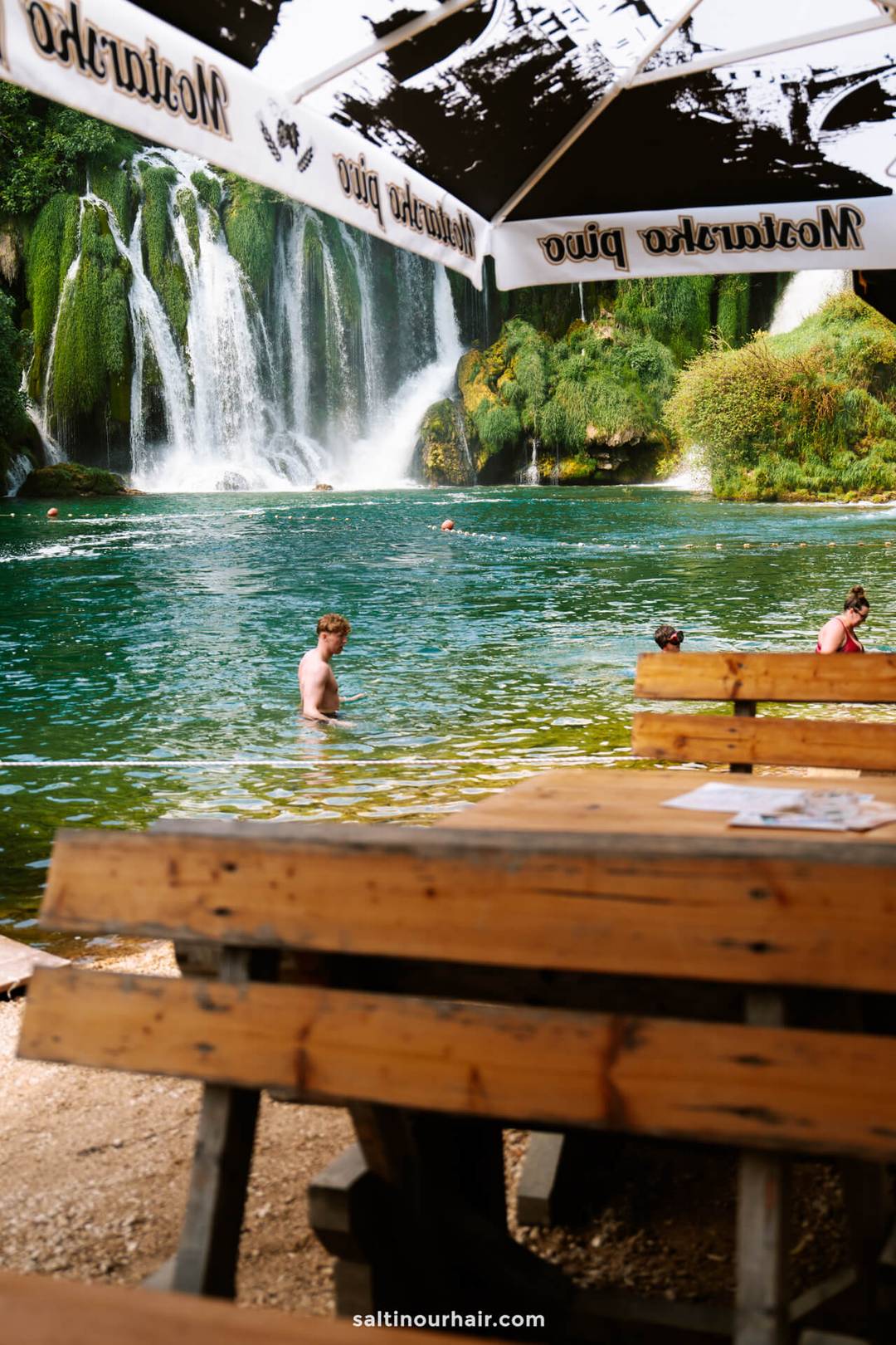 Swimmers in the turquoise pool at Kravica Waterfalls, with a Mostarsko Pivo umbrella and wooden picnic table in the foreground.