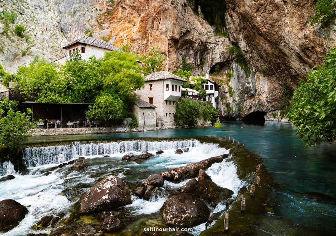 Stone buildings sit beside a river with a small waterfall, surrounded by greenery and steep rocky cliffs&mdash;one of the many picturesque things to do in Mostar, Bosnia.